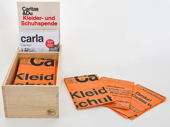 In einer kleinen Holzbox befinden sich die zusammengefaltenen orangen Kleidersäcke. 