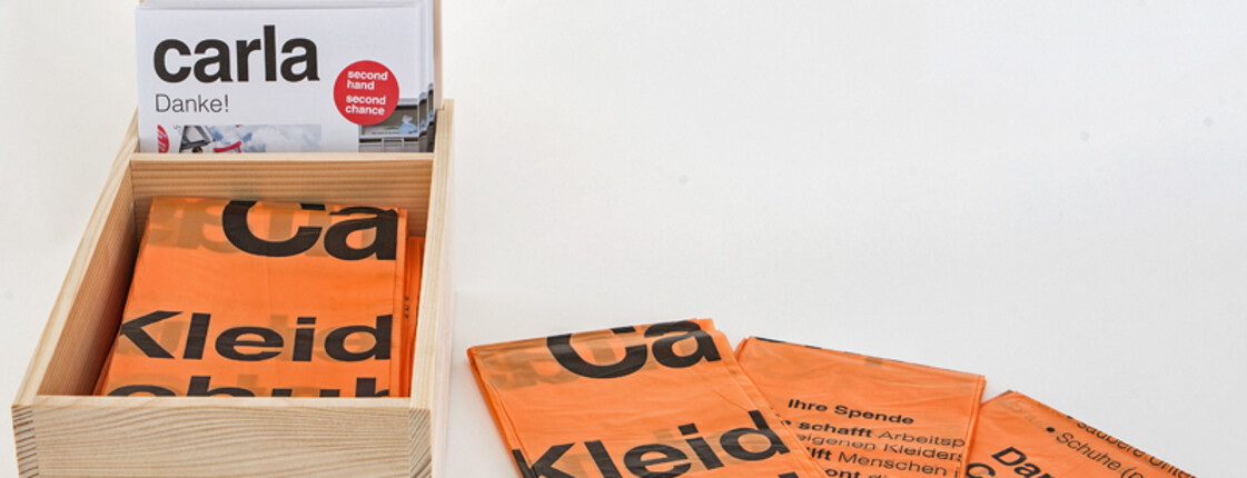 In einer kleinen Holzbox befinden sich die zusammengefaltenen orangen Kleidersäcke. 