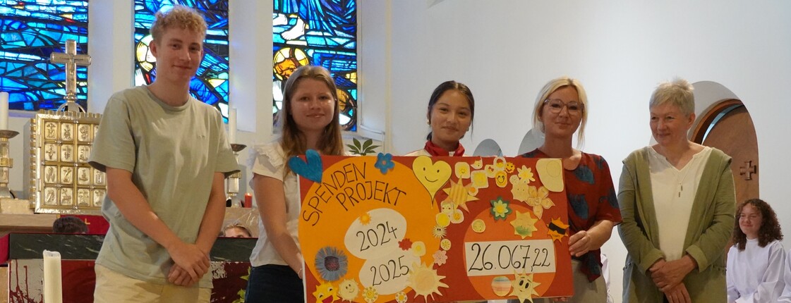 In einer Kirch präsentieren vier Jugendliche sowie eine Erwachsene einen selbst gemachten Spendenscheck in Höhe von rund 26.000 Euro.