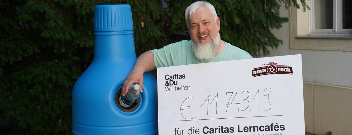 Caritas Direktor Norbert Filipitsch steht mit einem Spendenscheck neben einem Pfandaufsteller in Form einer blauen PET-Flasche.