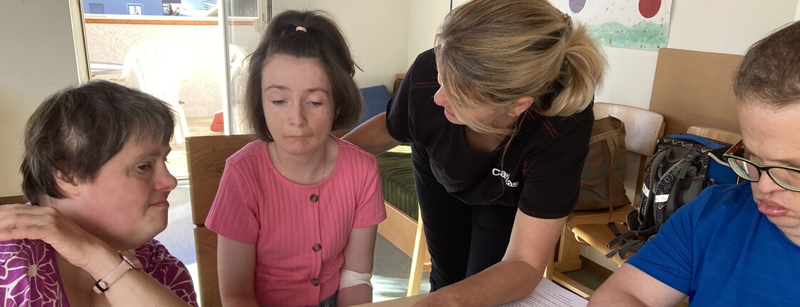 Drei Menschen mit Behinderungen sitzen an einem Tisch, auf dem ein aufgeschlagenes Fotoalbum liegt. Eine Caritas Mitarbeiterin zeigt auf eines der Fotos.