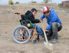 Baumplfanzaktion in Armenien: Ein Kind, das im Rollstuhl sitzt, und ein junger Erwachsener graben ein Loch in die Erde, um dann den Setzling darin zu pflanzen. Das Kind ist vormals in schwarz gekleidet, der junge Mann in blau. 
