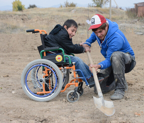 Baumplfanzaktion in Armenien: Ein Kind, das im Rollstuhl sitzt, und ein junger Erwachsener graben ein Loch in die Erde, um dann den Setzling darin zu pflanzen. Das Kind ist vormals in schwarz gekleidet, der junge Mann in blau. 