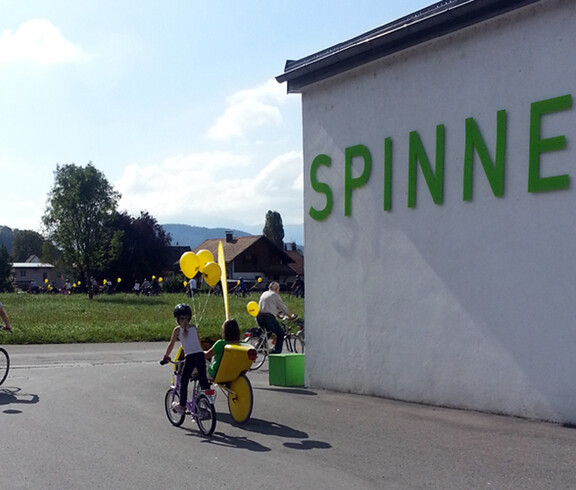 Ein Kind fährt mit einem Fahrrad auf Asphalt. Ans Rad wurde ein gelber Luftballon gebunden. Rechts zwei Meter daneben steht eine weiße Wand, auf der "Die Spinnerei" mit grünen Buchstaben steht. 