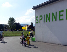 Ein Kind fährt mit einem Fahrrad auf Asphalt. Ans Rad wurde ein gelber Luftballon gebunden. Rechts zwei Meter daneben steht eine weiße Wand, auf der "Die Spinnerei" mit grünen Buchstaben steht. 