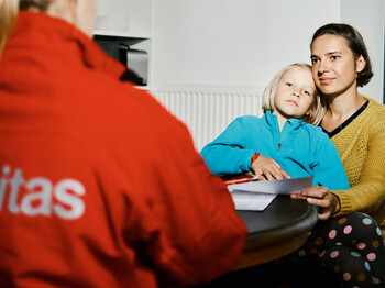 Eine Frau in roter Caritas-Jacke sitzt vor einer Frau mit kurzen Haaren, die ihre Tochter auf dem Schoß hat.