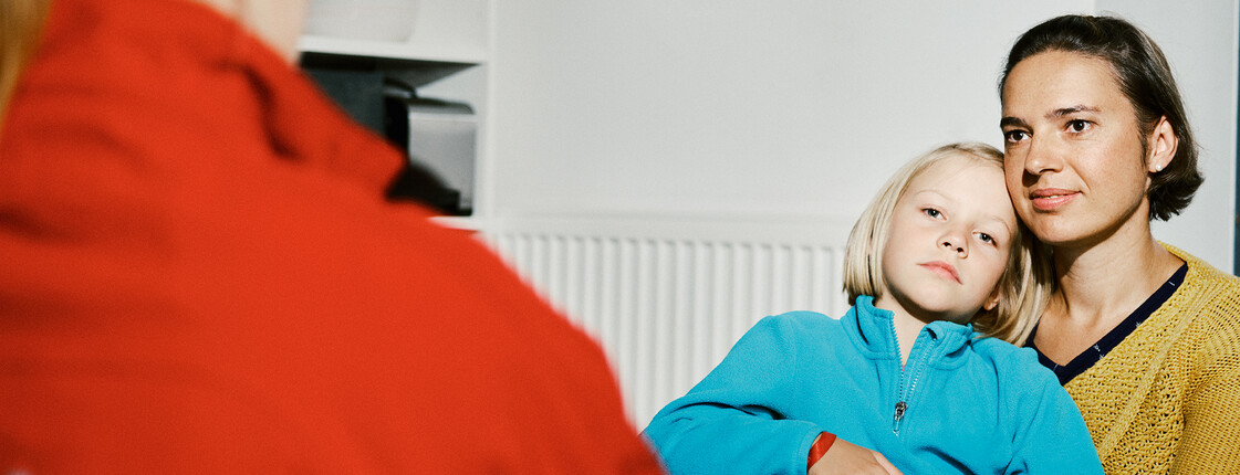 Eine Frau in roter Caritas-Jacke sitzt vor einer Frau mit kurzen Haaren, die ihre Tochter auf dem Schoß hat.