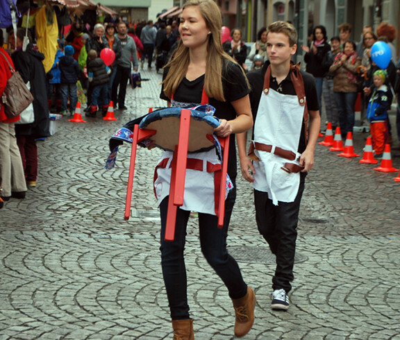 Eine junge Erwachsene und ein junger Erwachsener tragen schwarze Kleidung mit einer weißen Schürtze. Sie präsentieren diese Kreation in der feldkircher Innnenstadt Passanten am carla Reuse Day. 