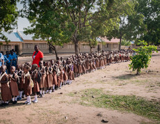 Viele kleine Kinder in Schuluniform stehen in einer langen Schlange vor einem Schulgebäude, neben ihnen sind ein paar Lehrer*innen zu sehen.