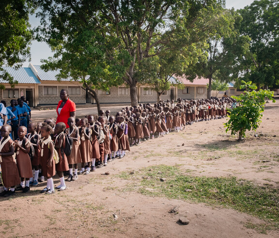 Viele kleine Kinder in Schuluniform stehen in einer langen Schlange vor einem Schulgebäude, neben ihnen sind ein paar Lehrer*innen zu sehen.