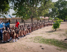 Viele kleine Kinder in Schuluniform stehen in einer langen Schlange vor einem Schulgebäude, neben ihnen sind ein paar Lehrer*innen zu sehen.