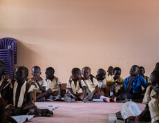 Kleine Kinder in Schuluniformen sitzen auf dem Boden in einem Raum mit rosaroten Wänden.