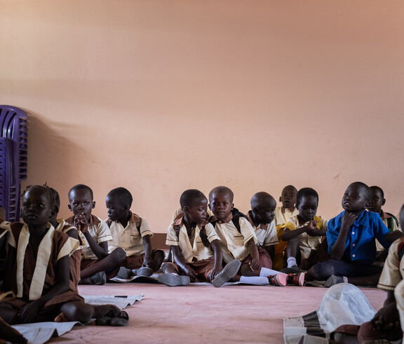 Kleine Kinder in Schuluniformen sitzen auf dem Boden in einem Raum mit rosaroten Wänden.