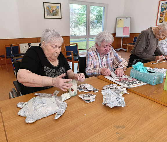 Mehrere Bewohner*innen gestalten mit Papierstreifen und Kleister Papiermaché-Figuren an einem Tisch im Aufenthaltsraum.