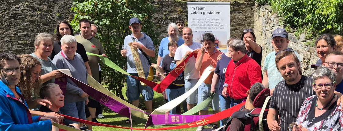 Gruppe von Teilnehmenden hält bunte Bänder im Kreis; gemeinsames Symbol für Pfingsten und Verbindung zwischen Gott und Mensch im Freien