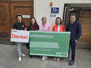 Fünf Personen stehen vor einem Gebäude und halten gemeinsam ein großes grünes Spendenscheck‑Plakat mit der Aufschrift „Danke! Spendenscheck 1.000 Euro“.