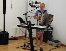 Musiker spielt Akkordeon neben Keyboard und Mikrofon bei Feier im Pflegewohnhaus, Caritas Pflege Rollup im Hintergrund.