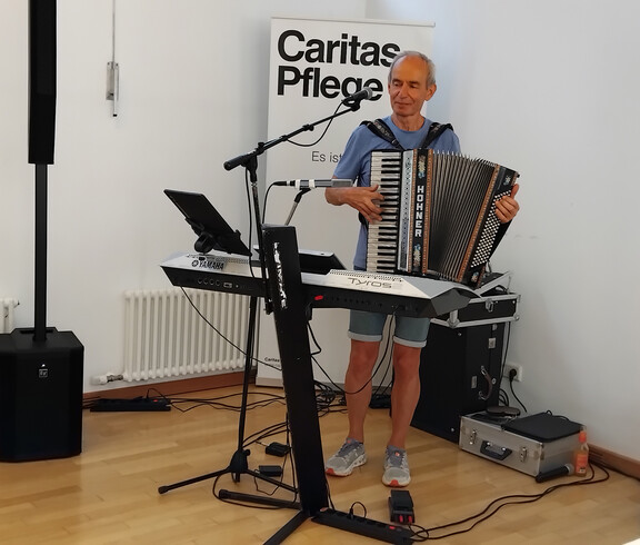 Musiker spielt Akkordeon neben Keyboard und Mikrofon bei Feier im Pflegewohnhaus, Caritas Pflege Rollup im Hintergrund.
