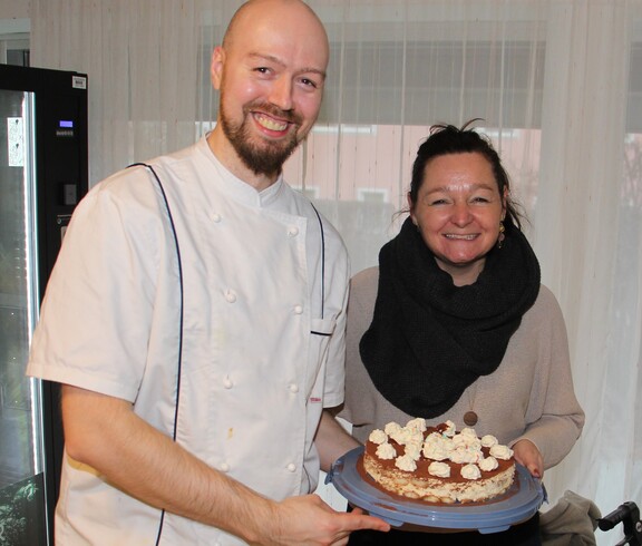 Die Hausleiterin und der Küchenchef präsentieren gemeinsam die Geburtstagstorte und lächeln in die Kamera.