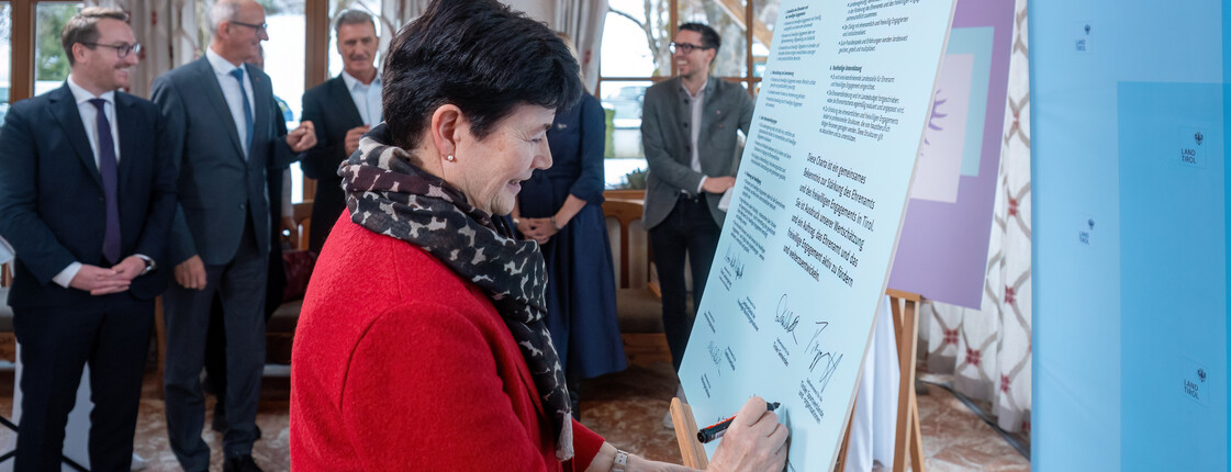 Eine Frau, die eine rote Jacke und einen Schal trägt, setzt ihre Unterschrift unter eine Charta, die auf einer Staffelei steht.