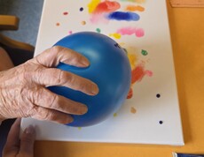 Eine Bewohnerin des Pflegewohnhauses arbeitet mit einem blauen Luftballon und bunten Farben, um durch Abdrücke ein kreatives und farbenfrohes Kunstwerk auf einer Leinwand zu gestalten