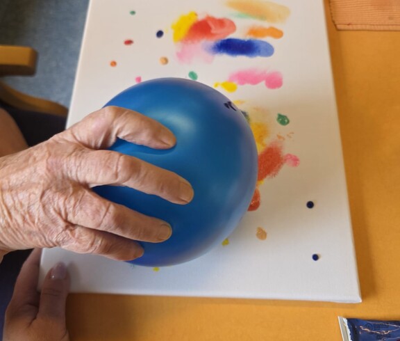 Eine Bewohnerin des Pflegewohnhauses arbeitet mit einem blauen Luftballon und bunten Farben, um durch Abdrücke ein kreatives und farbenfrohes Kunstwerk auf einer Leinwand zu gestalten