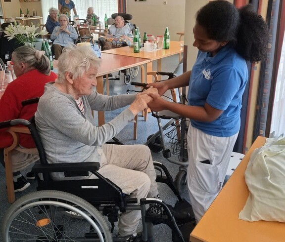 Eine Betreuungsperson hält die Hände einer sitzenden Bewohnerin und begleitet deren Bewegungen, während im Hintergrund weitere Personen gemeinsam Zeit im Aufenthaltsraum verbringen.