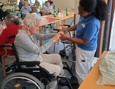 Eine Betreuungsperson hält die Hände einer sitzenden Bewohnerin und begleitet deren Bewegungen, während im Hintergrund weitere Personen gemeinsam Zeit im Aufenthaltsraum verbringen.