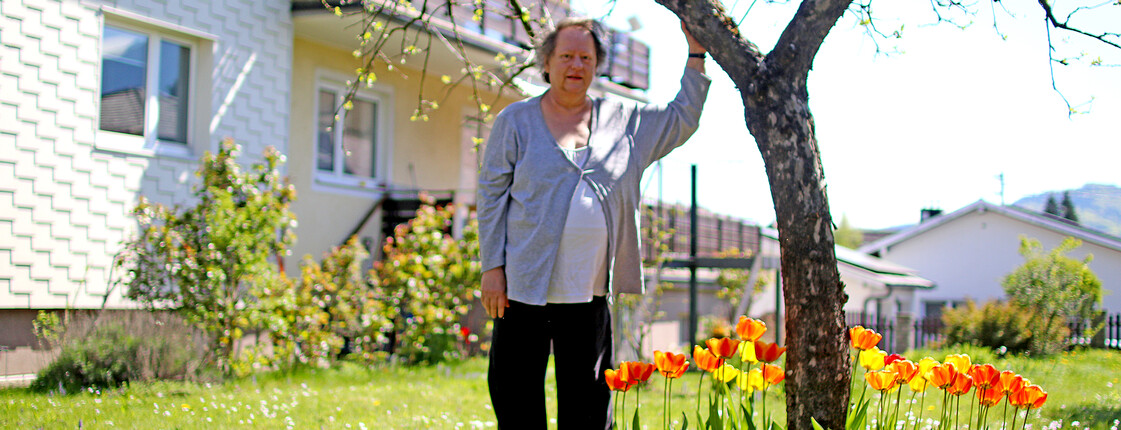 Eine Frau steht unter einem Baum in einem Garten neben einen prächtigen Blumenbeet. Dahinter ein Wohnhaus.