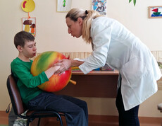 Eine Therapeutin unterstützt ein Kind im Kinderdorf Gomel bei einer medizinischen Übung mit einem großen Ball, um seine Motorik und Beweglichkeit zu fördern.