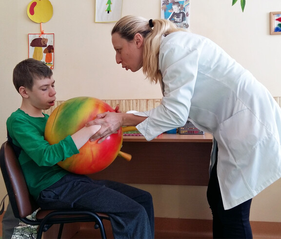 Eine Therapeutin unterstützt ein Kind im Kinderdorf Gomel bei einer medizinischen Übung mit einem großen Ball, um seine Motorik und Beweglichkeit zu fördern.