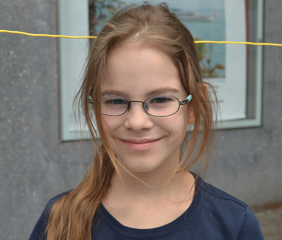 Ein junges Mädchen im Volksschulalter blickt in die Kamera. Sie hat dunkelblonde lange Haare und trägt eine Brille. Sie hat ein dunkelblaues Oberteil an. 