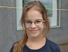 Ein junges Mädchen im Volksschulalter blickt in die Kamera. Sie hat dunkelblonde lange Haare und trägt eine Brille. Sie hat ein dunkelblaues Oberteil an. 