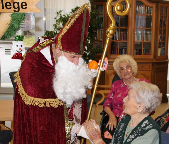 Nikolaus überreicht einer sitzenden Bewohnerin ein kleines Geschenk. Beide lächeln, im Hintergrund ist eine weitere Bewohnerin zu sehen.