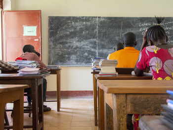 Zu sehen ist der Rücken von Erwachsenen, die in einem Klassenzimmer sitzen und auf eine Tafel schauen.