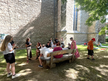 Menschen sitzen und stehen an einem Tisch im Schatten neben einer Pfarrkirche und kommen miteinander ins Gespräch.