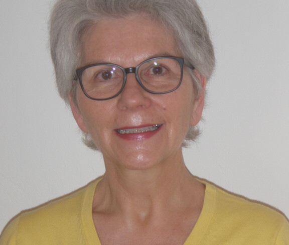 Das Foto zeigt eine Frau mittleren Alters. Sie trägt ein gelbes Oberteil, roten Lippenstift und eine schwarze Brille.