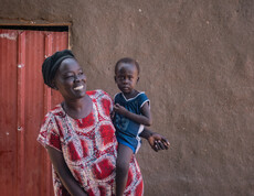 Eine junge Frau lacht und hält ihr Kleinkind auf ihrer Hüfte. Sie trägt bunte traditionelle Kleidung und steht vor einem Lehmhaus.