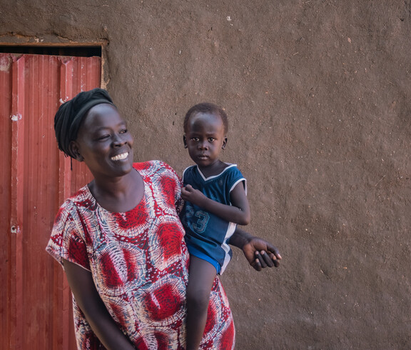 Eine junge Frau lacht und hält ihr Kleinkind auf ihrer Hüfte. Sie trägt bunte traditionelle Kleidung und steht vor einem Lehmhaus.