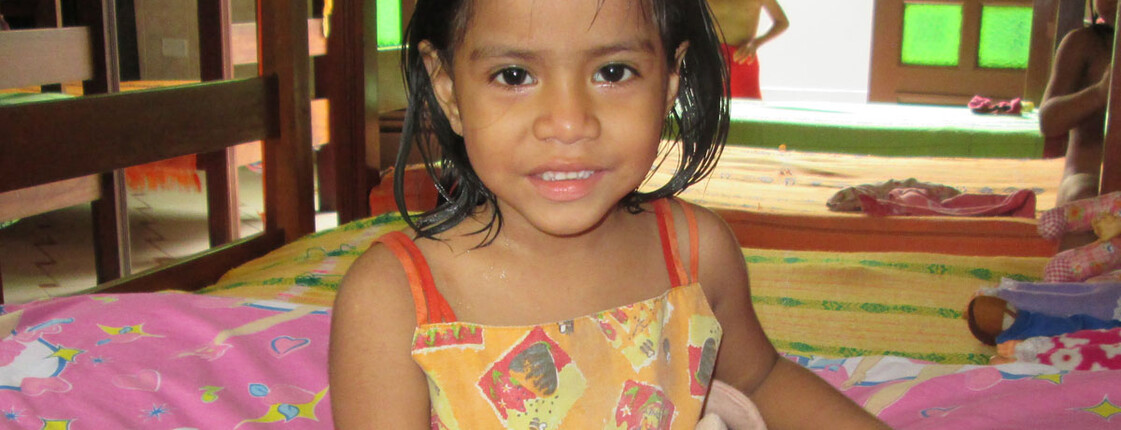Ein kleines Mädchen in buntem Kleid sitzt lächelnd auf einem Bett im Mädchenheim El Huambrillo in Iquitos, Peru; im Hintergrund sind Etagenbetten und weitere Kinder zu sehen.
