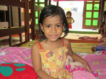 Ein kleines Mädchen in buntem Kleid sitzt lächelnd auf einem Bett im Mädchenheim El Huambrillo in Iquitos, Peru; im Hintergrund sind Etagenbetten und weitere Kinder zu sehen.