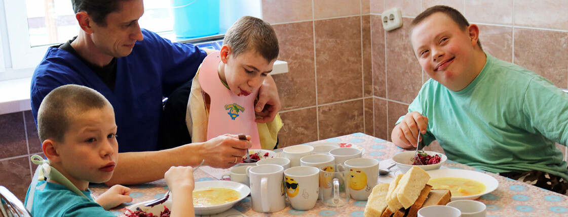 Kinder im Kinderdorf Gomel nehmen gemeinsam mit einem Betreuer ihre Mahlzeiten ein, wobei einige Kinder unterstützt werden.