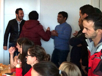 Über zehn Menschen stehen in einem kleinen Esszimmer. Dabei auffallend: Alle Frauen sitzen am Tisch während die Männer im Raum stehen und am Diskutieren sind. Die abgebildeten Personen sind schlicht bekleidet und tragen eher dünklere Farben. 