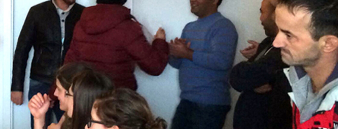 Über zehn Menschen stehen in einem kleinen Esszimmer. Dabei auffallend: Alle Frauen sitzen am Tisch während die Männer im Raum stehen und am Diskutieren sind. Die abgebildeten Personen sind schlicht bekleidet und tragen eher dünklere Farben. 