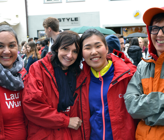 Vier Caritas Mitarbeiterinnen nahmen bei einem Laufwettbewerb teil. Sie stehen in der feldkircher Innenstadt zu einem Gruppnfoto zusammen. Drei tragen eine rote Regenjacke. Die andere eine orange graue. Im Hintergrund sind etliche Menschen in einer Masse versammelt. 