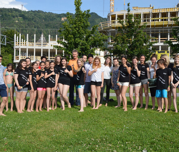 Jugendliche stehen mit einem erwachsenen Herrn auf einer Wiese. Sie sind leicht bekleidet. Der Großteil trägt schwarze T-Shirts. 