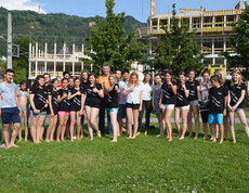 Jugendliche stehen mit einem erwachsenen Herrn auf einer Wiese. Sie sind leicht bekleidet. Der Großteil trägt schwarze T-Shirts. 