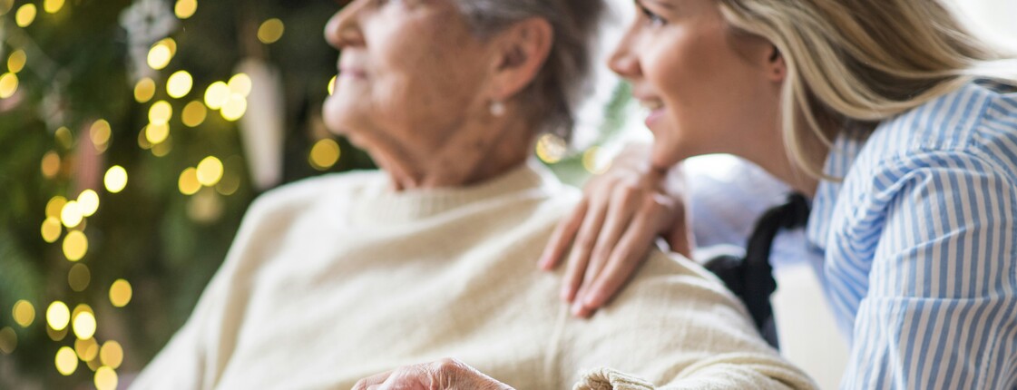 Eine junge Frau sitzt neben einer älteren Dame, hält einfühlsam ihre Hand und erklärt ihr etwas mit freundlichem Gesichtsausdruck.
