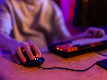 Close-up einer Hand auf Tastatur und Maus, beleuchtet vom violetten Licht eines PC-Bildschirms.