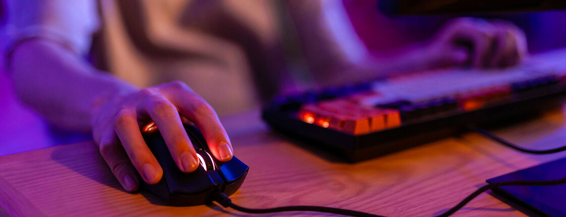 Close-up einer Hand auf Tastatur und Maus, beleuchtet vom violetten Licht eines PC-Bildschirms.